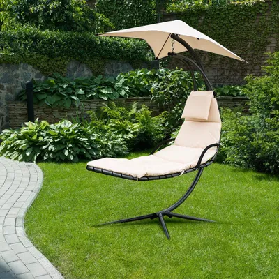 A beige chaise lounge with a sunshade is set up on a green lawn in a garden, with lush green plants and a stone wall in the background.