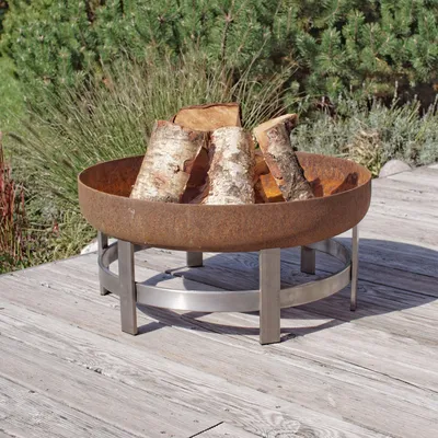 A rusty metal fire pit filled with logs sits on a wooden deck, with greenery in the background.