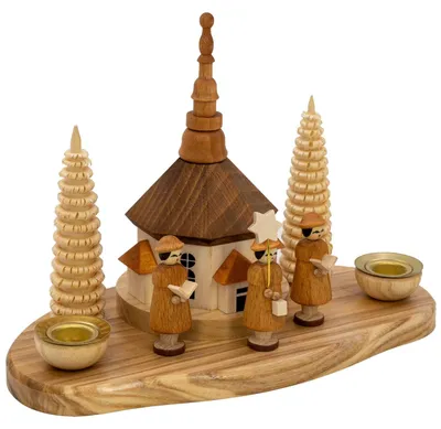 Auf einem hölzernen Tablett stehen drei geschnitzte Figuren in braunen Gewändern mit einem Stern und Notenblättern vor einer kleinen Kirche flankiert von zwei spiralförmigen Bäumen und zwei Teelichthaltern.