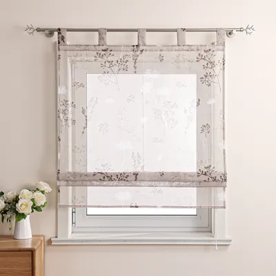 A sheer Roman blind with a floral pattern is shown hanging over a window. A vase of white roses sits on a wooden surface to the left of the window.