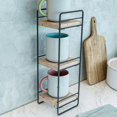 A three-tiered shelf unit holding three mugs, placed on a countertop next to a wooden cutting board.