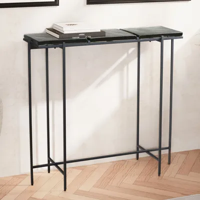 A narrow console table with a black metal frame and two dark, segmented glass top surfaces, placed against a white wall with a herringbone wooden floor.