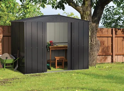 A dark gray metal garden shed with its doors open, revealing a workbench and stool inside. A green wheelbarrow filled with soil and a shovel are to the left of the shed. A wooden fence and a large tree are in the background.