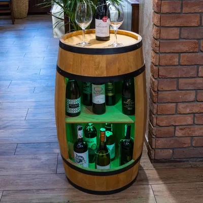 A wooden barrel-shaped wine cabinet with shelves and bottles, illuminated by green LED lights, placed in a room with wooden floors and a brick wall.