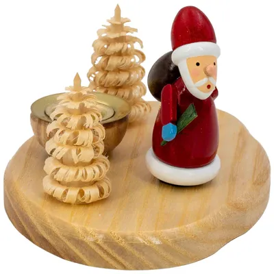 Ein hölzerner Kerzenhalter zeigt einen kleinen Weihnachtsmann in rotem Gewand mit einem grünen Zweig in der Hand neben zwei kunstvoll geschnitzten Bäumen auf einer runden Holzplatte