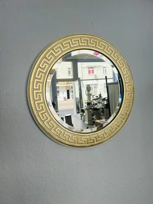A round mirror with a gold ornate frame hangs on a light gray wall. The mirror reflects a shop interior.