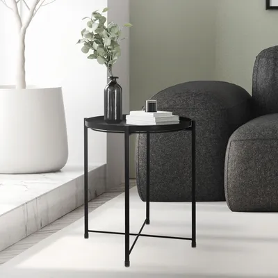 A black metal side table with a removable tray is placed next to a dark gray sofa in a modern living room. A vase with green leaves and a stack of books are on the table. A large white planter with a decorative tree is to the left.
