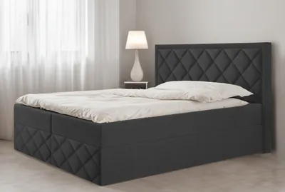 A dark gray upholstered box spring bed with a diamond-stitched headboard and base, made up with white bedding, is shown in a bedroom setting with sheer white curtains and a white table lamp on a nightstand.