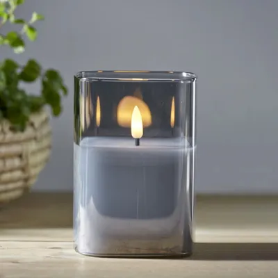 A lit LED candle in a glass container sits on a wooden surface, with a plant in a basket in the background.