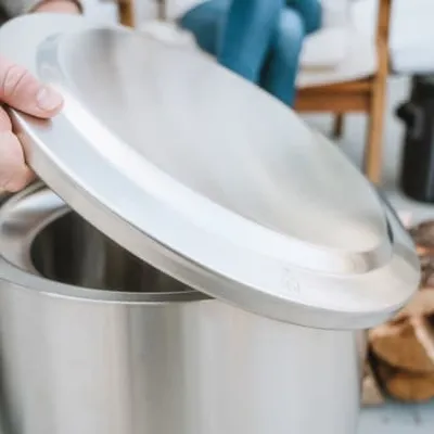 A hand is lifting the lid off a stainless steel fire pit. In the background, people are sitting on chairs and there is a stack of firewood.