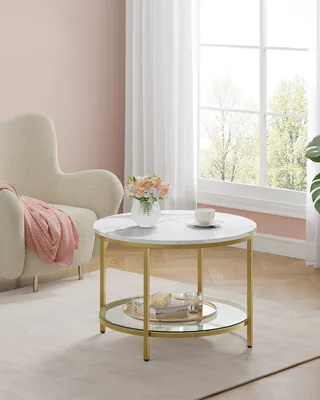 A round coffee table with a marble top and gold legs, placed in a living room setting next to an armchair.