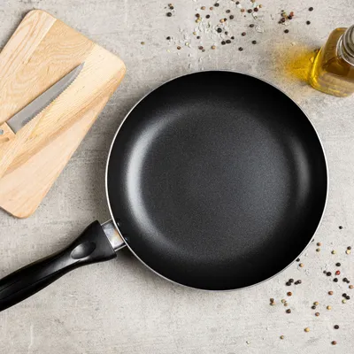 A frying pan, cutting board with a knife, and a bottle of oil are arranged on a textured gray surface, with scattered peppercorns.