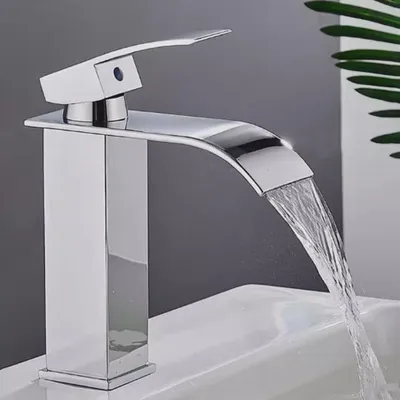 A modern chrome waterfall faucet is shown in use over a white sink, with a green leaf in the background.
