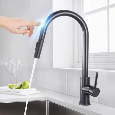 A hand is touching a black kitchen faucet, activating the water flow. The faucet is modern and sleek, with a pull-down sprayer. There are wine glasses and lime slices on the counter in the background.