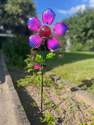 A solar garden stake shaped like a flower with purple and pink petals and a red glass center, placed in a garden bed.