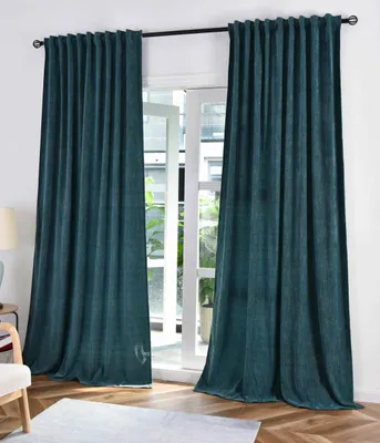 Two teal curtains are hung on a curtain rod in front of a window. The curtains have a subtle textured pattern. The floor is made of wooden planks, and a light-colored rug is visible in the foreground.