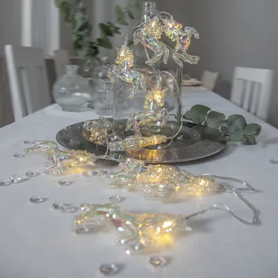 A string of unicorn-shaped LED lights is arranged on a white tablecloth, with a glass bottle and other decorative elements in the background.