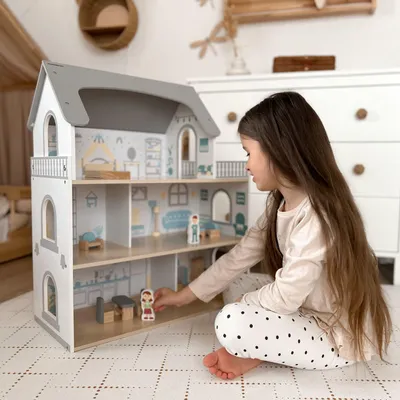 A young girl is playing with a wooden dollhouse in a room.