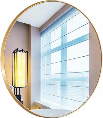 A round mirror with a gold frame reflecting a room with a window and a lamp.