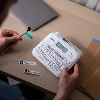 A person is using a Brother P-touch label maker to label a cable connected to a laptop.