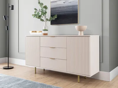 A modern sideboard with decorative fluting and gold legs, placed in a well-lit room with a floor lamp and artwork.