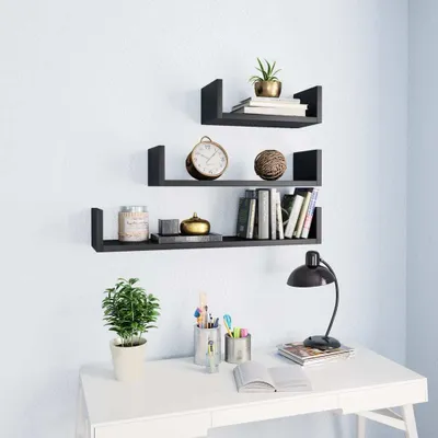 Three black U-shaped shelves mounted on a white wall, decorated with books, a clock, a plant, and decorative items. Below, a white desk with a lamp, a potted plant, and pens in a holder.