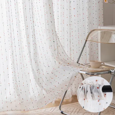 A close-up view of sheer curtains with colorful pom-poms, draped over a clear chair with a cup of tea on a tray.
