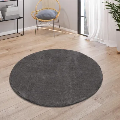 A round, dark gray rug is placed on a wooden floor in a room with a chair and a shelf in the background.