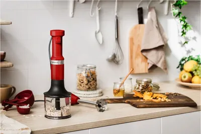 A red KitchenAid immersion blender is shown on a kitchen counter with ingredients and kitchen utensils in the background.