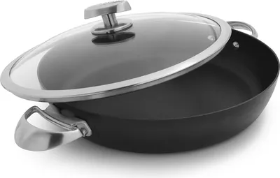 A black frying pan with a glass lid and silver handles is shown from a slightly elevated side angle against a white background.