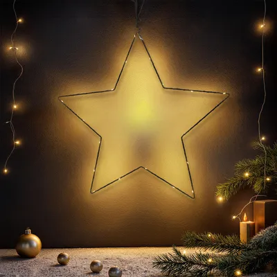 A star-shaped light decoration is hung on a dark wall, with Christmas ornaments and pine branches on the floor below.