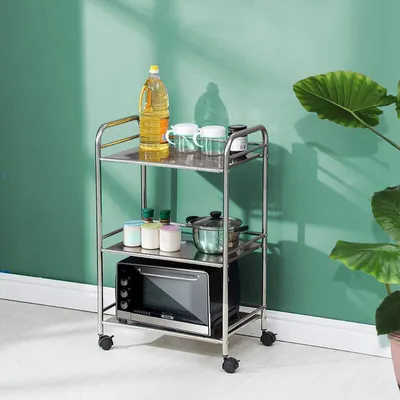 A stainless steel kitchen cart with three shelves, holding various kitchen items like oil, containers, and a microwave, placed against a green wall.
