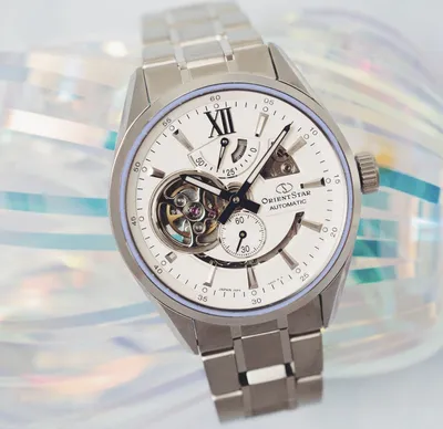 A close-up shot of a silver Orient Star automatic watch with a white dial and a skeletonized movement.