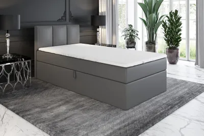 A grey boxspring bed with a white mattress and topper is shown in a bedroom setting with plants and a nightstand.
