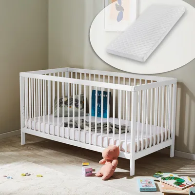 A white baby crib with a mattress and pillows inside, placed on a wooden floor with a rug and toys nearby. A close-up of a mattress is shown in an inset circle at the top.