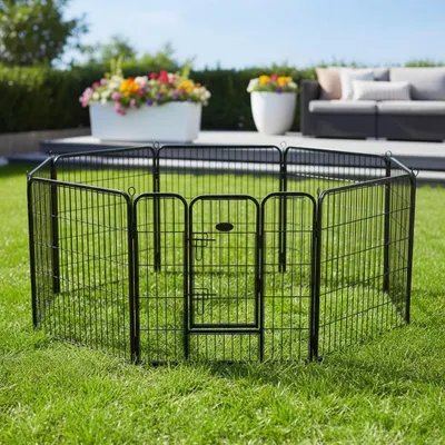 A black metal pet playpen is set up on a green lawn in a backyard setting. In the background, there are flower pots with colorful flowers, a patio, and outdoor furniture.