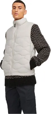 A man wearing a beige puffer vest over a black and gray patterned sweater and dark pants.