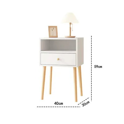 A white nightstand with a drawer and an open shelf, placed on a white background. It has wooden legs and a wooden drawer knob. A small framed picture and a lamp are on top of the nightstand.