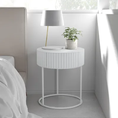 A white round bedside table with a plant and a lamp on it, next to a bed.