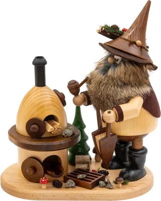 Ein Erzwichtel mit spitzem Hut und buschigem Bart steht neben einem kleinen Holzofen und hält einen Hammer in der Hand während um ihn herum kleine Holzscheite und Tannenzapfen liegen