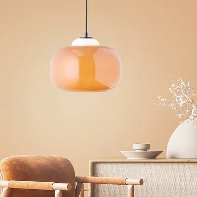 A pendant lamp with an orange glass shade hangs above a table with a vase and a chair in the foreground.