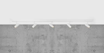 A white track light fixture with five adjustable spotlights is mounted on a white ceiling above a textured concrete wall.