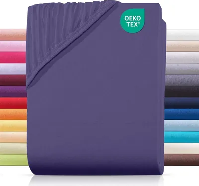 A folded purple fitted sheet with a small green OEKO-TEX logo in the top right corner. A stack of sheets in various colors is visible on the left and right sides of the image.