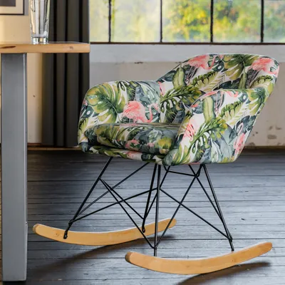 A rocking chair with a tropical flamingo and leaf pattern is placed in a room with large windows and wooden floors.