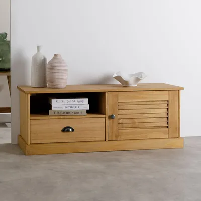 A wooden TV lowboard with one door and one drawer, placed on a concrete floor against a white wall. Decorative vases and books are on top and inside the unit.