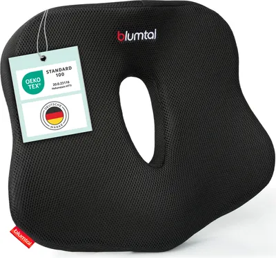A black ergonomic seat cushion with a cut-out center, featuring certification tags attached.