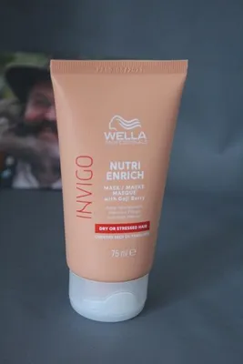 A tube of Wella Invigo Nutri Enrich hair mask is shown against a gray background, with a blurred image of a person in the background.