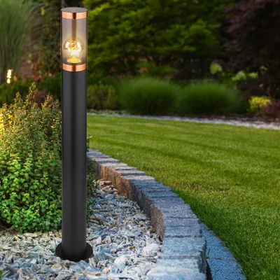 A black bollard light with a copper top and glass shade is illuminated in a garden at dusk, next to a curved stone border and lush greenery.