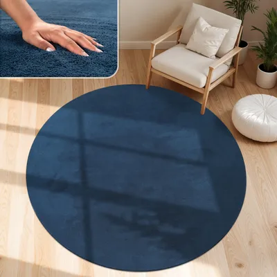 A round, dark blue rug is placed on a light wooden floor in a living room setting. A chair and a pouf are visible in the background.