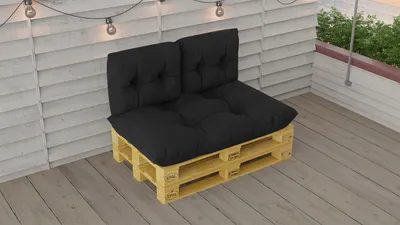 A black pallet cushion set is placed on a wooden deck against a white slatted wall, with string lights overhead.